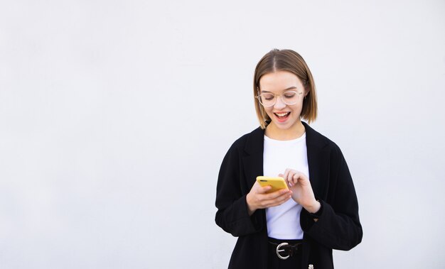 Vrouw die een jasje en glazen dragen die een smartphone en glimlachen gebruiken, geïsoleerd op wit
