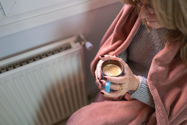 Vrouw die een kopje koffie drinkt naast een radiator die koud wordt in de winter