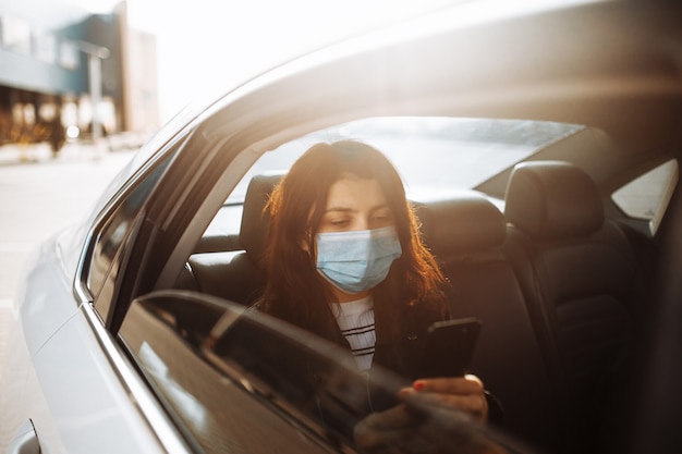 Vrouw die een medisch steriel masker draagt in een taxiauto op een achterbank die uit venster kijkt