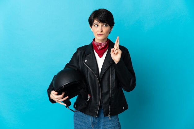 Vrouw die een motorhelm houdt die op blauwe muur met vingers kruist en het beste wenst