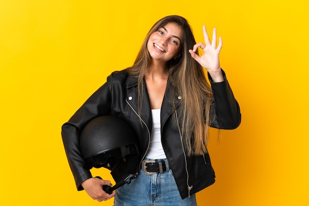 Vrouw die een motorhelm houdt die op geel wordt geïsoleerd dat ok teken met vingers toont