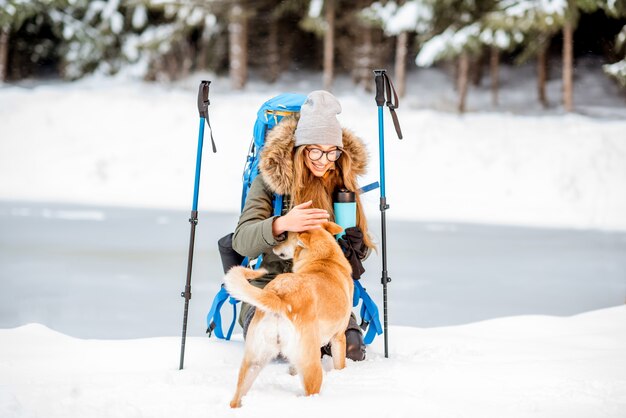 Vrouw die een pauze heeft tijdens de winterwandeling en haar hond aait in de besneeuwde bergen bij het meer en het bos