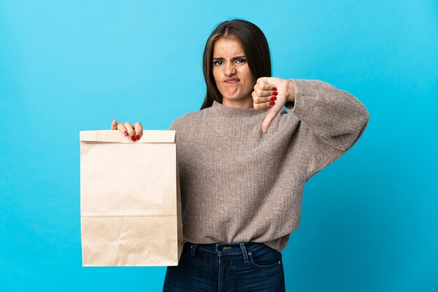 Vrouw die een zak afhaalmaaltijden neemt die op blauwe muur wordt geïsoleerd die duim met negatieve uitdrukking toont