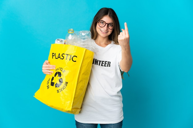 Vrouw die een zak vol flessen houdt om te recyclen geïsoleerd op blauwe muur die komend gebaar doet