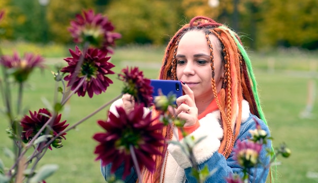 Vrouw die foto's maakt op smartphone in park Positieve jonge vrouwelijke hipster met lange dreadlocks in vrijetijdskleding die bloemen op mobiele telefoon in park fotografeert