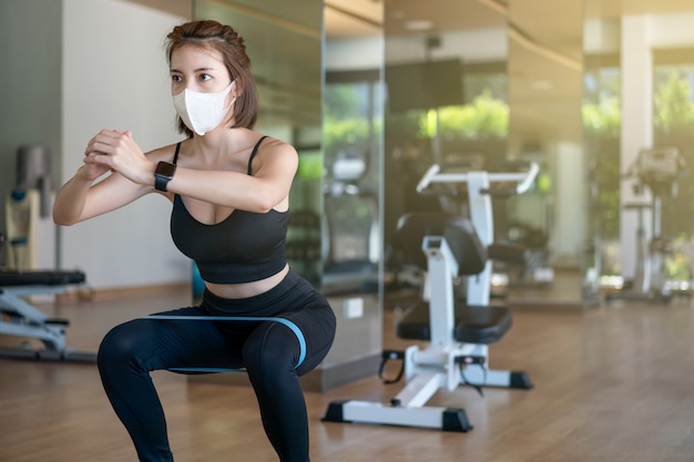 Vrouw die gezichtsmasker draagt, hurkzit met buit apparatuur uitrekkende riem in een fitnesscentrum. tijdens de pandemie van het coronavirus.