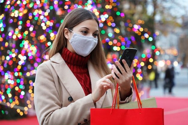 Vrouw die gezichtsmasker draagt op straat met boodschappentassen en slimme telefoon voor online aankopen met kleurrijke kerstboomverlichting