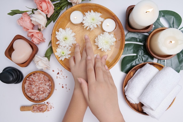 Vrouw die haar handen in een kom met water en bloemen drenkt, kuur en product voor vrouwelijke voeten en handkuuroord, massagekiezel, geparfumeerd bloemenwater en kaarsen, ontspanning. Plat leggen. bovenaanzicht.