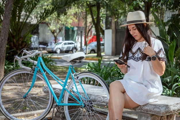Vrouw die haar mobiele telefoon controleert terwijl ze in het park zit met een fiets naast haar in een jurk