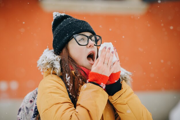 Vrouw die haar neus met een weefsel openlucht in de winter blaast. Jonge vrouw die ziek met griep in een de winterdag wordt