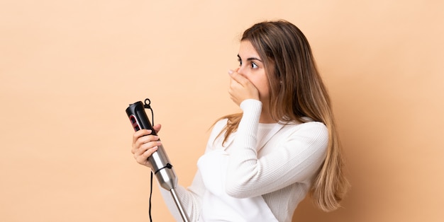 Vrouw die handmixer over geïsoleerde achtergrond gebruikt die mond behandelt en aan de kant kijkt