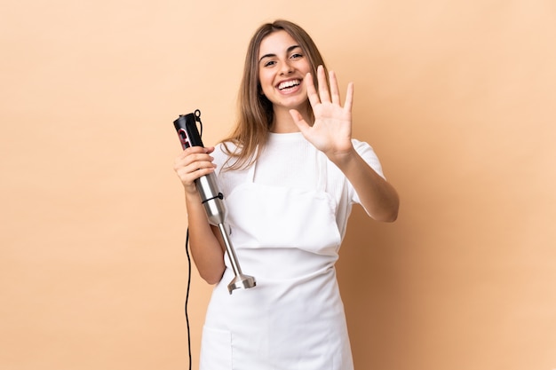 Vrouw die handmixer over geïsoleerde achtergrond gebruikt die vijf met vingers telt