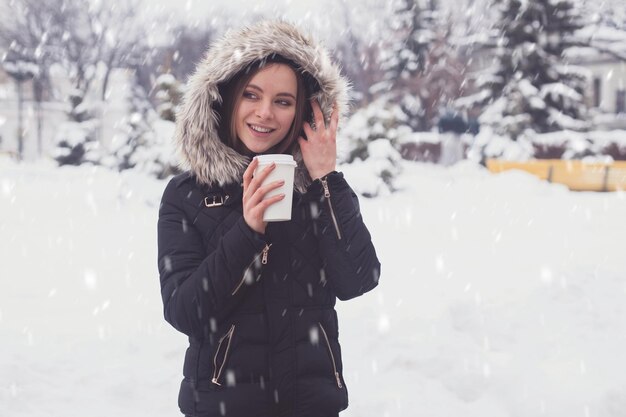 Vrouw die hete koffie of thee drinkt uit een mok onder sneeuwvlokken in de winter