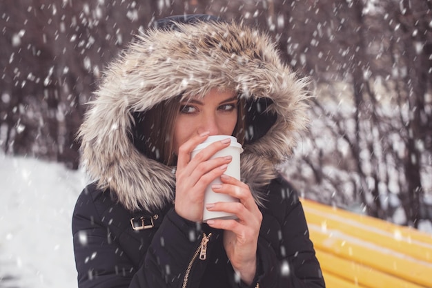 Vrouw die hete koffie of thee drinkt uit een mok onder sneeuwvlokken in de winter