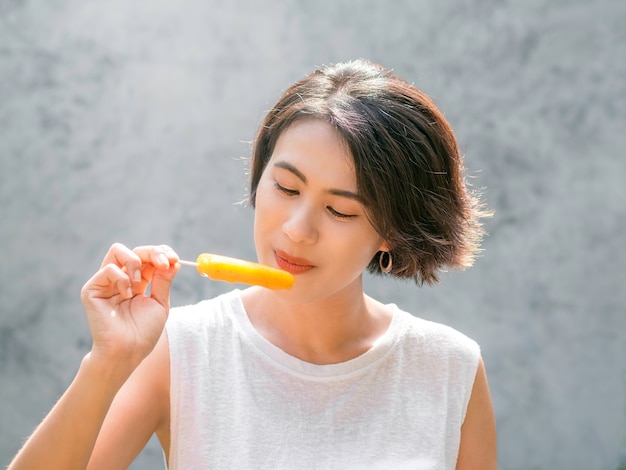 Vrouw die ijslollys eet. Gelukkige mooie Aziatische vrouw die een toevallig wit mouwloos overhemd draagt dat gele ijslolly houdt, in openlucht. Glimlachend vrouwtje genieten van ijslolly in de zomer.