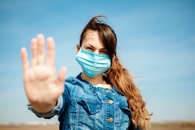 Vrouw die in beschermend steriel medisch masker op haar gezicht camera in openlucht bekijkt. Hand stopbord. Pandemisch coronavirus concept.