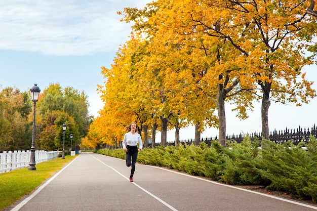 Vrouw die in de herfstbos loopt. Jong volwassen meisje joggen in herfstkleuren, camera kijken en brede glimlach.