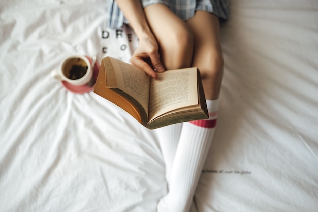 Vrouw die in een wit bed ligt, thuis thee drinkt en boek leest.