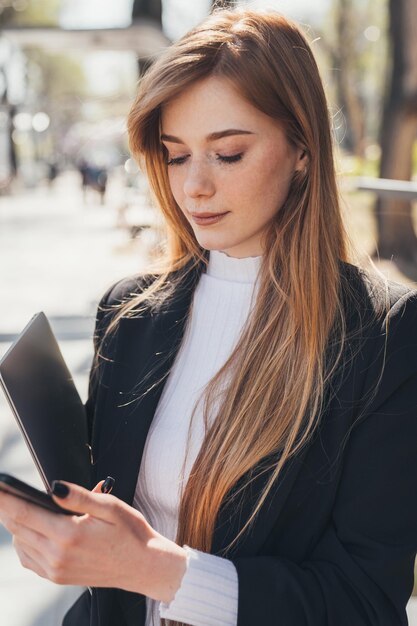 Foto vrouw die in het park staat te wachten op haar vriendin die te laat is met de telefoon om haar te bellen te laat op het werk te zijn moderne apparaten vasthoudend