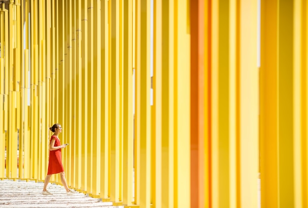 Vrouw die in rode kleding op de moderne gele achtergrond van de de bouwmuur loopt. Algemeen plan met abstracte geometrische compositie