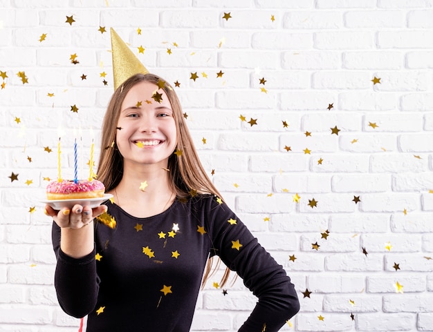 Vrouw die in verjaardagshoed een doughnut met kaarsen met gouden confettien houdt die rond vallen
