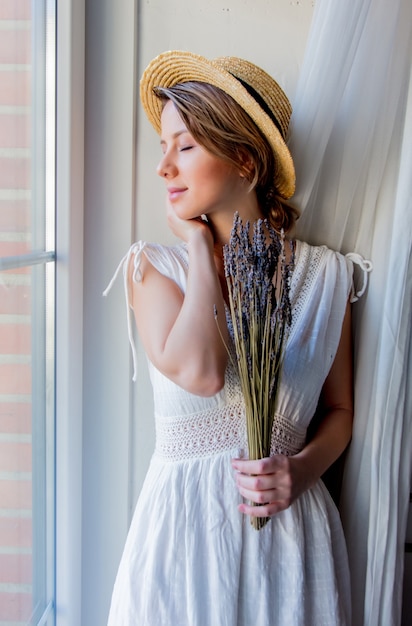 Vrouw die in witte kleding lavendel in handen houdt en dichtbij venster blijft. Afbeelding in hoge sleutelstijl