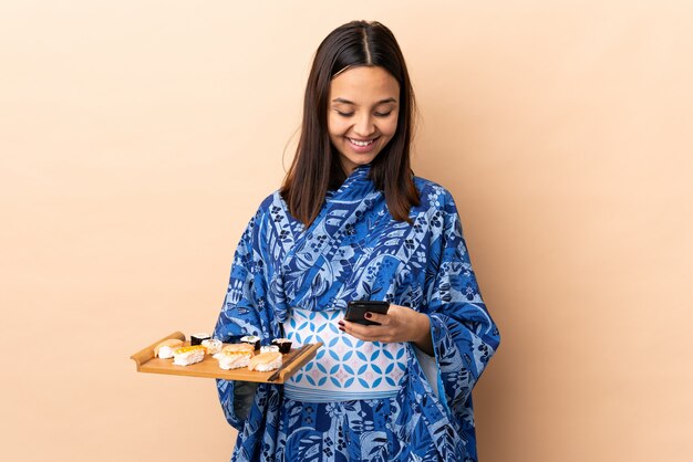 Vrouw die kimono draagt en sushi over geïsoleerde achtergrond houdt die een bericht met mobiel verzendt