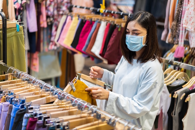 Vrouw die kleren kiest bij winkelcomplex en medisch masker draagt voor preventie van coronavirus