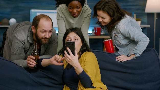 Vrouw die komische video op de telefoon laat zien aan haar multiraciale vrienden die samen socializen tijdens het nachtfeest. Groep mensen van gemengd ras die op de bank zitten, bier drinken, chillen in de woonkamer