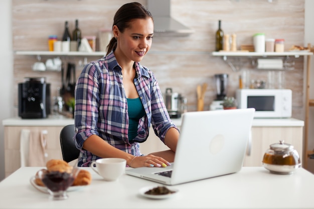 Vrouw die lacht met behulp van laptop in de keuken in de ochtend met een kopje hete groene thee naast haar