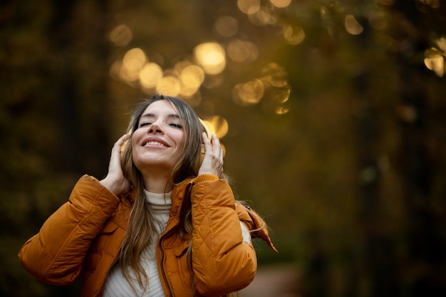 Vrouw die lacht met gesloten ogen en gezicht naar de hemel met koptelefoon en oranje jas in park bij zonsondergang