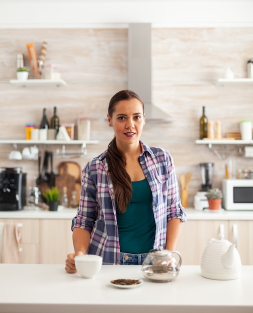 Vrouw die lacht naar de camera in de keuken met een kop hete thee tijdens het ontbijt