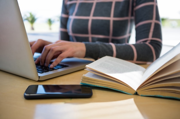 Vrouw die laptop met mobiele telefoon en roman op lijst met behulp van