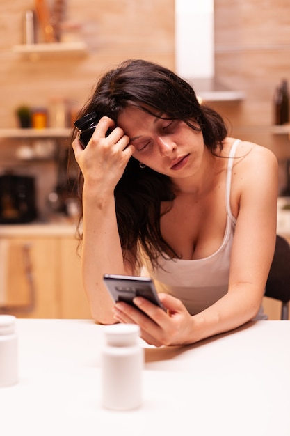 Vrouw die lijdt aan angst die op internet surft met behulp van smartphone in de keuken van het huis met pillen ervoor. Bezorgd onwel vrouw lijden aan migraine, depressie, ziekte en angst.