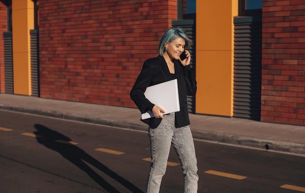 Vrouw die met collega's aan de telefoon praat terwijl ze door de straten van de stad loopt met een laptop in haar hand...