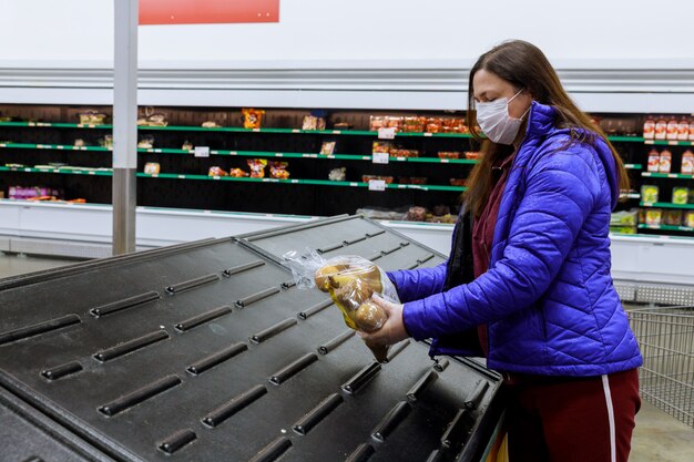 Vrouw die met gezichtsmasker laatste zak aardappel houdt bij supermarkt met lege planken.