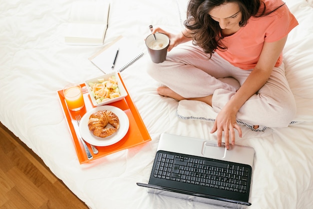 Vrouw die met haar laptopcomputer werkt en ontbijt. Ze is in haar slaapkamer.