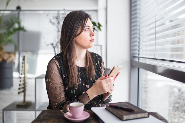 Vrouw die met mobiele telefoon door venster in koffiewinkel kijkt