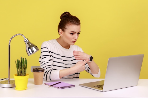 Vrouw die op kantoor zit en naar het horloge wijst terwijl ze naar het scherm van de laptop kijkt, uitgeput van overwerk