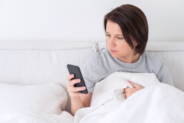 Vrouw die smartphone gebruiken aangezien zij in bed zit dat met dekbed wordt behandeld