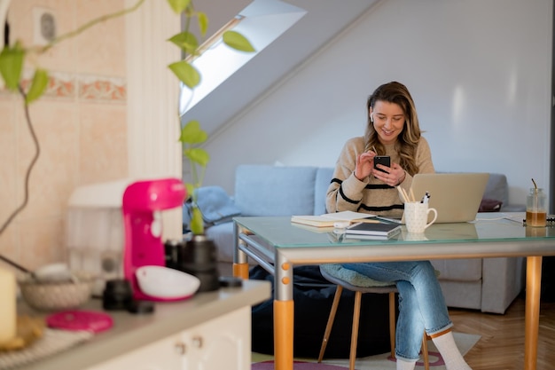 Vrouw die smartphone gebruikt terwijl ze thuis werkt en een laptop gebruikt