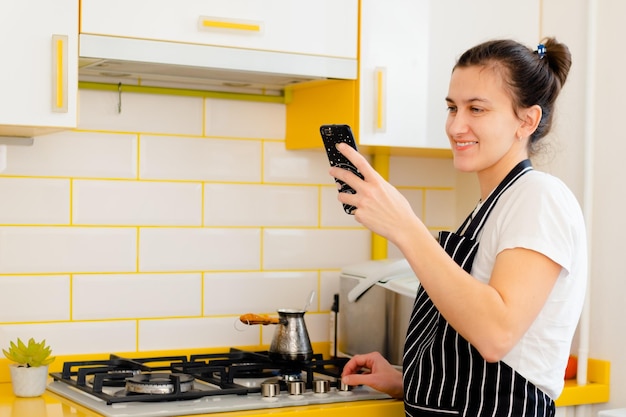 Vrouw die smartphone gebruikt voor videogesprekken en koffie zet in de keuken Concept happy morning leisure