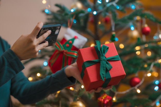 Vrouw die smartphone mobiel gebruikt, maakt een foto van een kerstcadeaudoos met een kerstboom op de achtergrond