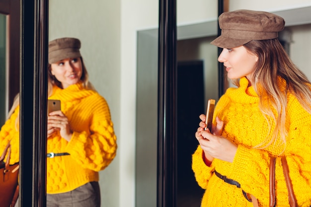 Vrouw die spiegel thuis het dragen van modieuze sweater en toebehoren bekijken en selfie nemen