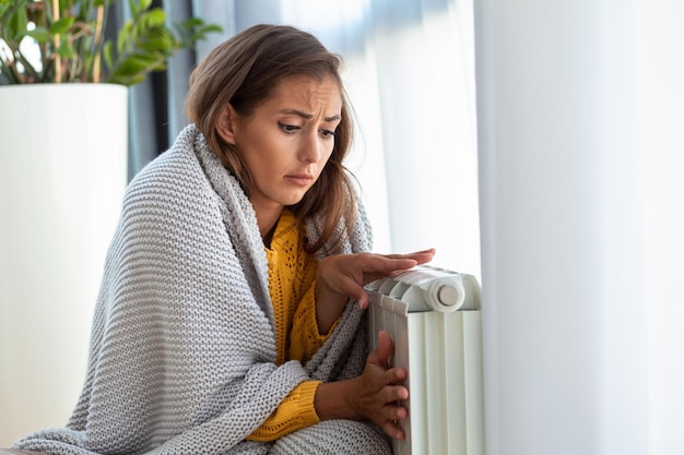 Vrouw die thuis bevriest terwijl ze bij de koude radiator zit Vrouw met een probleem met de verwarming in huis die het koud heeft Stroombeperking