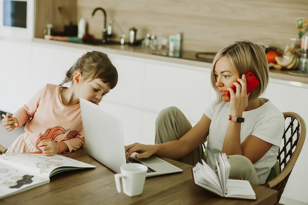 Vrouw die thuis werkt met haar dochter die in de buurt van moeder zit met laptop en telefonisch praat