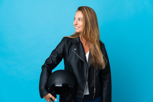 Vrouw die van middelbare leeftijd een motorhelm houdt die op blauwe muur wordt geïsoleerd die naar de kant kijkt en glimlacht