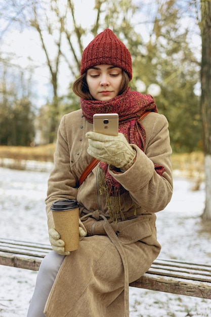 Vrouw die warme kleren draagt met een koffiekopje om mee te nemen die smartphone gebruikt en op een bankje in het park zit