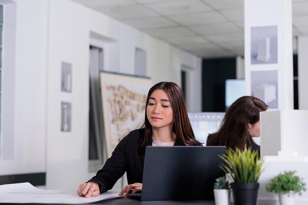 Vrouw die werkt met blauwdrukken in coworking space, aziatische werknemer in architectenbureau op werkplekbureau. Architect, vastgoedmedewerker, softwareontwikkelaar die projectplan leest