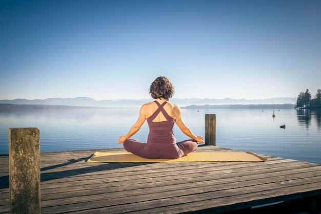 Vrouw die yoga beoefent op een oefenmat op de steiger boven het meer tegen een heldere lucht